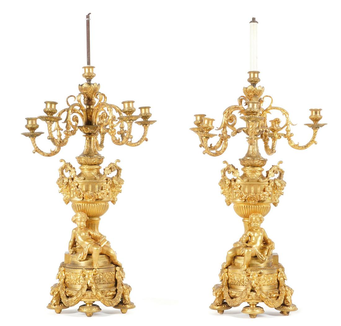 Pair French Gilt Bronze Figural Candelabra: Pair gilt bronze candelabra, each with six foliate scroll arms, electrified center fixture, urn form center support over figural putti, one reading and the other holding a painting of a tree. Base wit