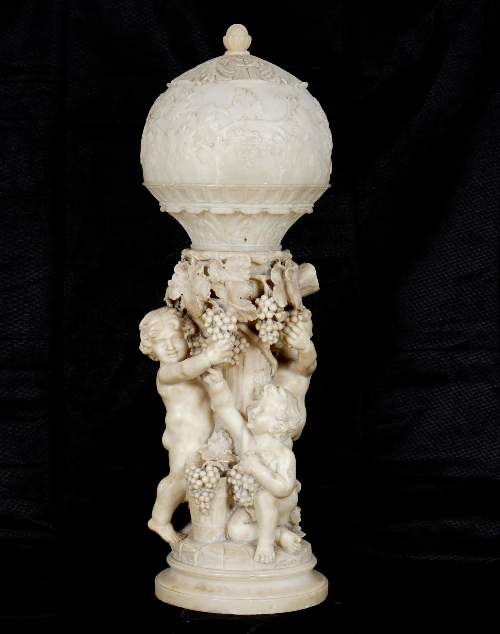 Large Carved Alabaster Bacchanalian Putti Lamp: Carved alabaster, three figural putti picking grapes off a grape vine on a column, foliate and grape vine motif domed shade, single socket fixture. Overall 27" h. x 11" base dia. Condition: Minor chip