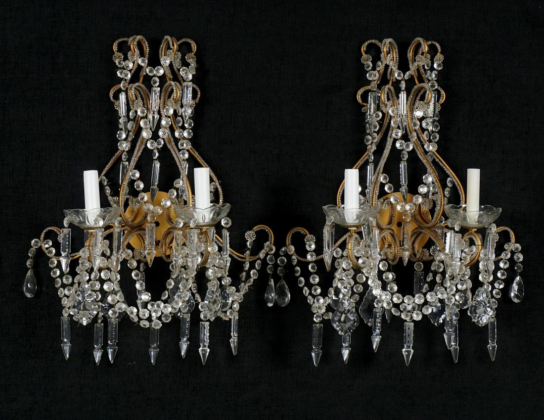 Pair Gilt Metal and Crystal Sconces: Gilt metal base and scroll arms, two electric lights, crystal prisms, drops, garland, and beading. Overall 23" h. x 17" x 8.75". Condition: The occasional chip or missing prism may be discovered. From