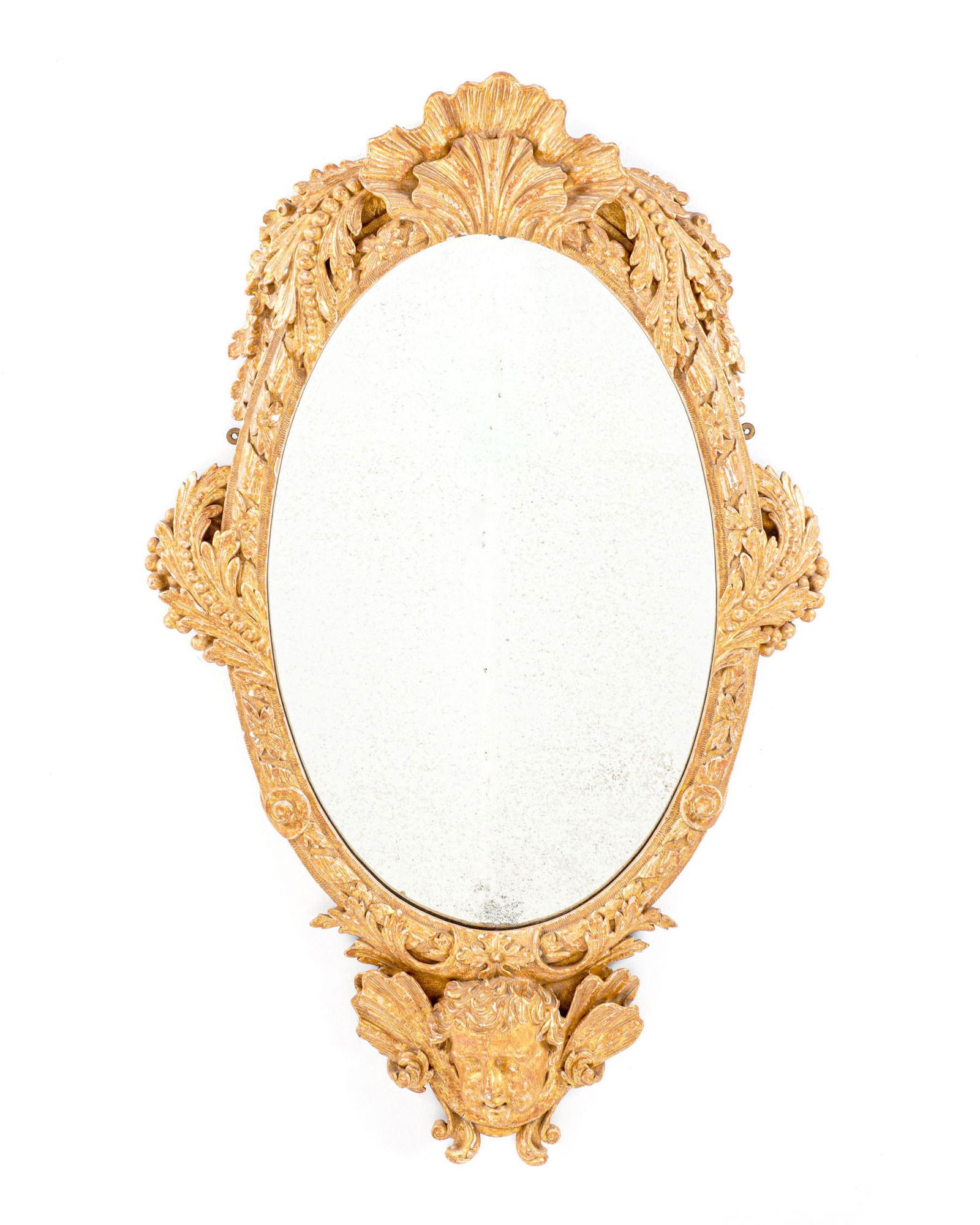 George II Manner of William Kent Gilt Wood Mirror: 18th Century, in the manner of William Kent, carved gilt wood frame comprising shell finial, foliate leaf and ribbon, lower cherub mask, oval center mirror. Overall 45.5" x 28". The family purchased i