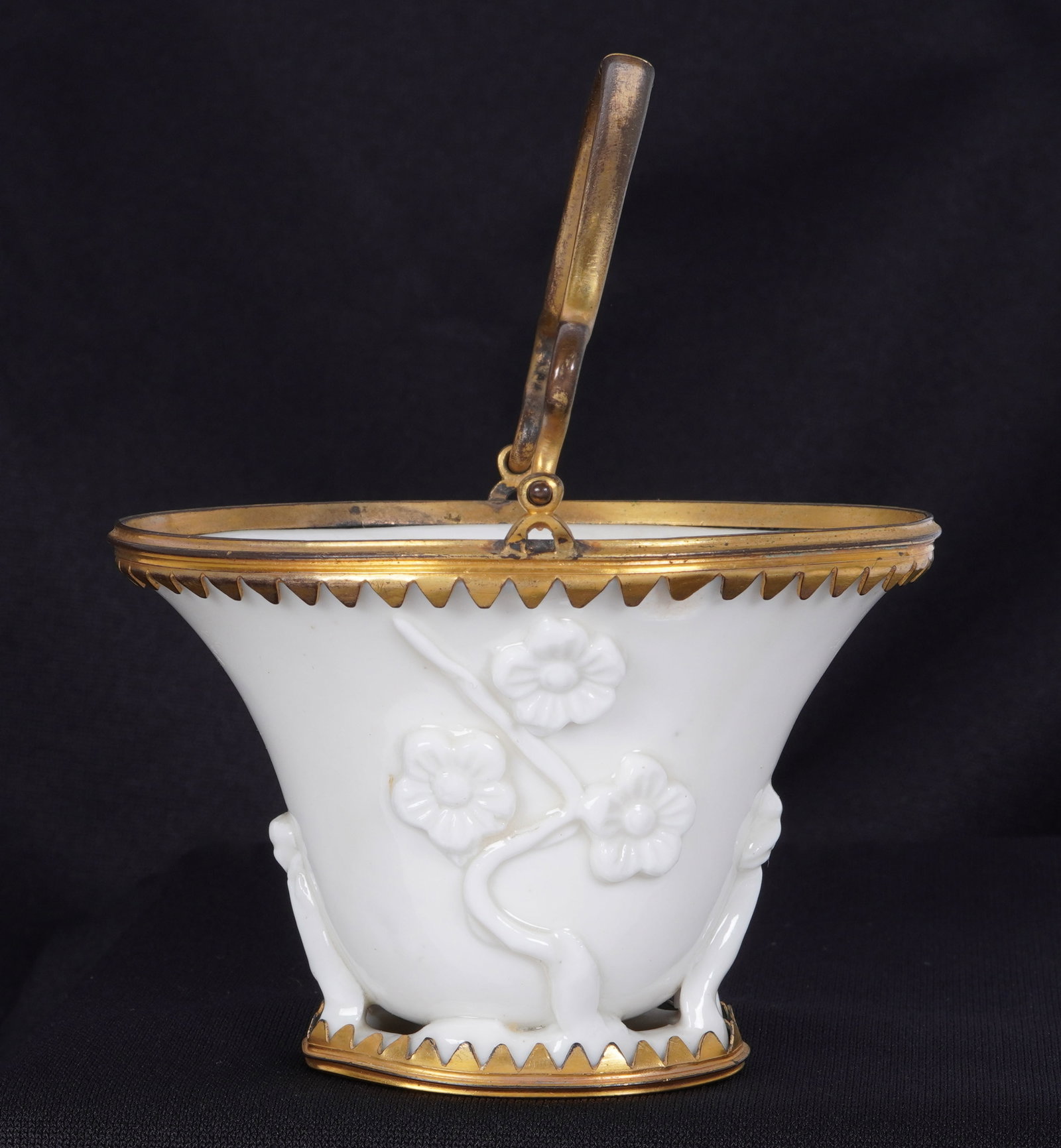 Chinese Blanc de Chine Brass Mounted Libation Cup: Floral decorated blanc de chine porcelain libation cup, brass mounted base, top rim with hinged handle, no visible markings. Overall 2.75" h. x 3.75" x 3.5". Condition: Nick to cup, missing one brass