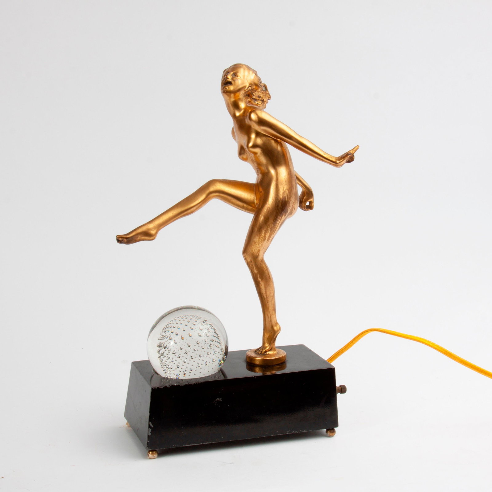 Figural Nude Art Deco Lamp: A figural nude Art Deco dancer lamp with gold patina. With a controlled-bubble glass orb set on a black-patinated metal base, outfitted with a turning switch. Measures 13 1/4" x 8 1/4" x 4".