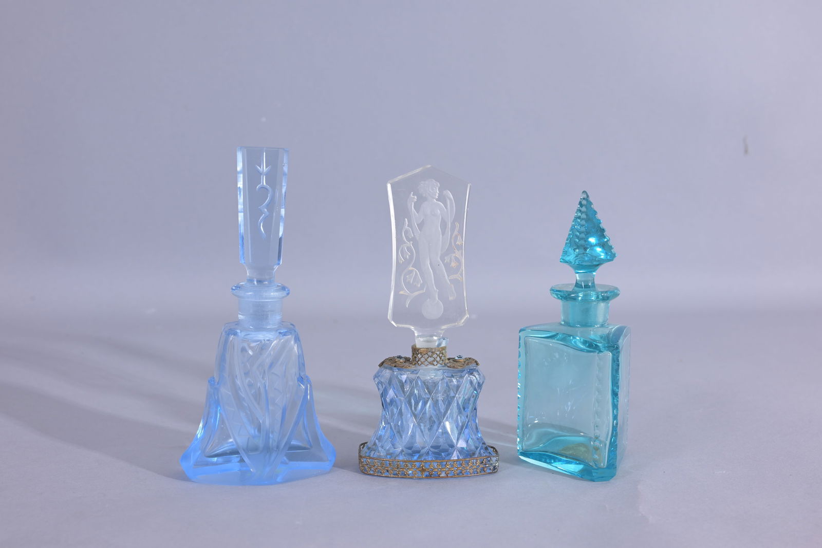 (3) Assorted Blue Czech Perfume Bottles: Medium size clear blue bottle. - Clear blue bottle, jeweled with gold lace like and blue gems. Clear stopper with nude lady standing on a sphere. - Irice Czech clear aqua blue perfume bottle, triangle