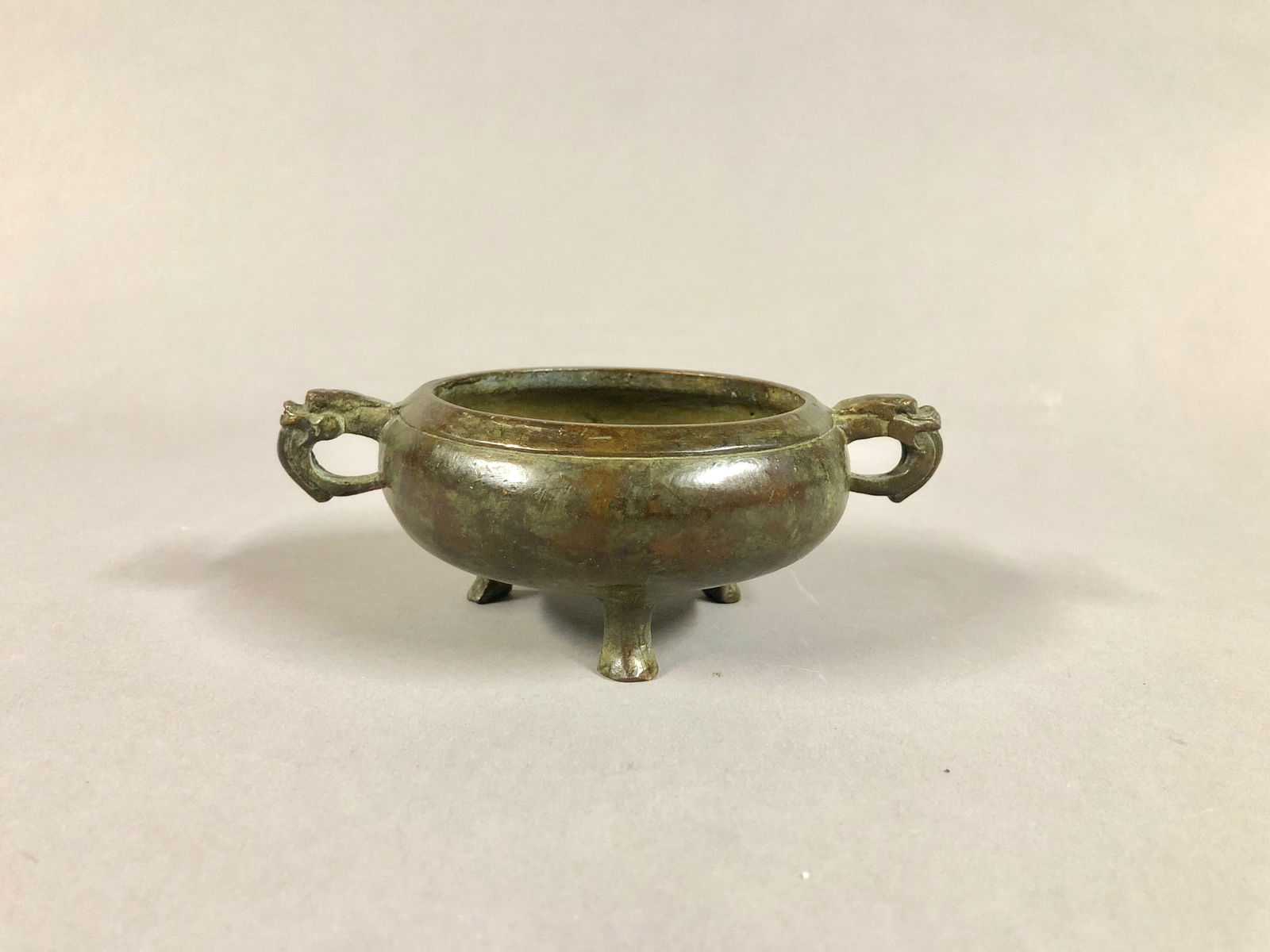 Small Chinese Bronze Incense Burner: A Small Chinese Bronze Incense Burner of plain form, with Dragon Head handles, resting on 3 feet, and having characters on its base. Dimensions: 1.75 H x 5 W in.