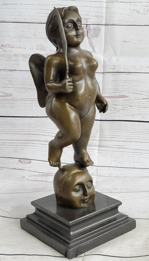 Standing Angel Bronze Sculpture: The plump angel stands nude walking upon a decollated head that smiles with chubby cheeks under the weight. She looks out at the beholder, bold with an amiable gentleness, holding a dagger upward in h