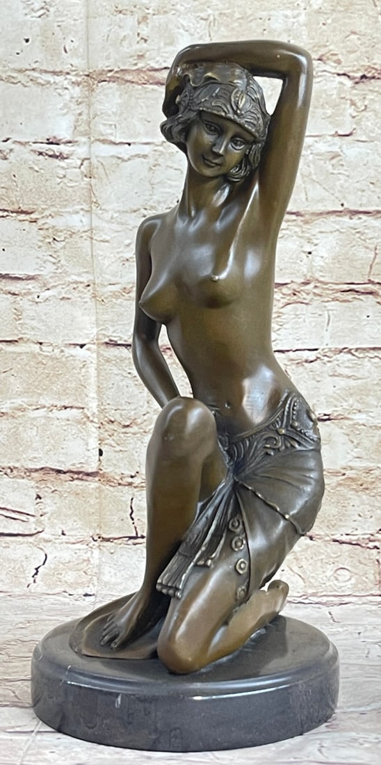 Nude Woman Kneels Bronze Statue: The nude beauty, adorned with only a translucent beaded skirt and headpiece, kneels on her left knee in an alluring pose. Her left-hand bends and crowns her head that faces down. This Bronze Sculpture