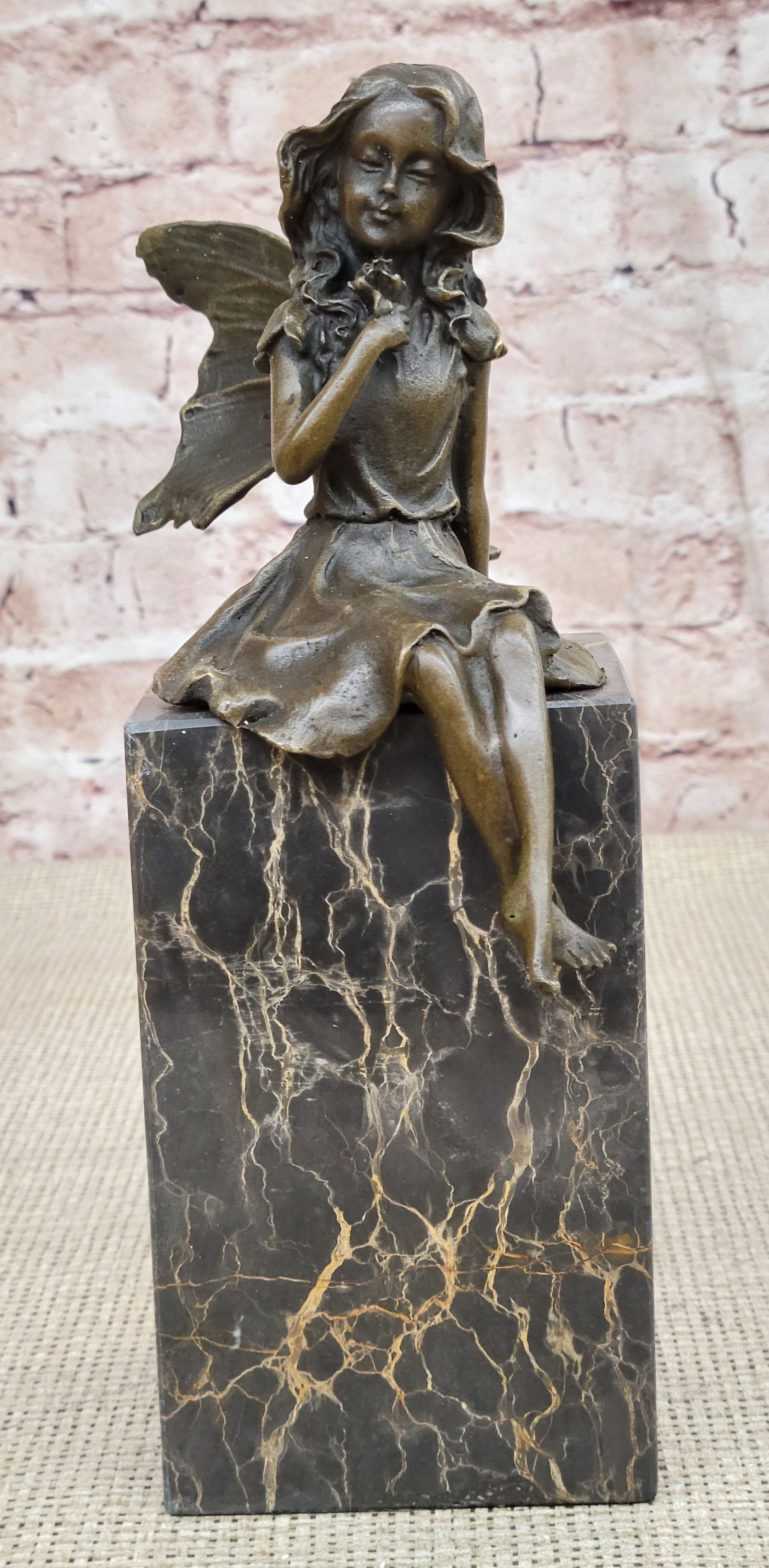 Angel Holding Flower Bronze Figurine: Daydreamers will fall in love with this daydreaming fairy. She sits on a rock and gazes out into the mystical land around her. She dangles her legs over the side and crosses her ankles. She is lulled