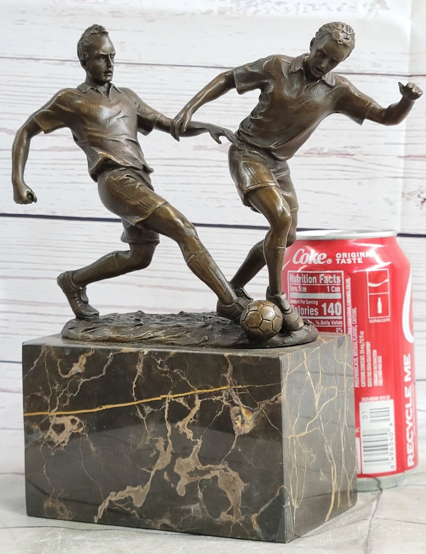 Two Soccer Player Bronze Sculpture: They are legends on the field. Two soccer players tumble down the grassy field, kicking, and dodging along the way. They are swift on their feet. One nears the goalpost and lunges one leg forward, pro