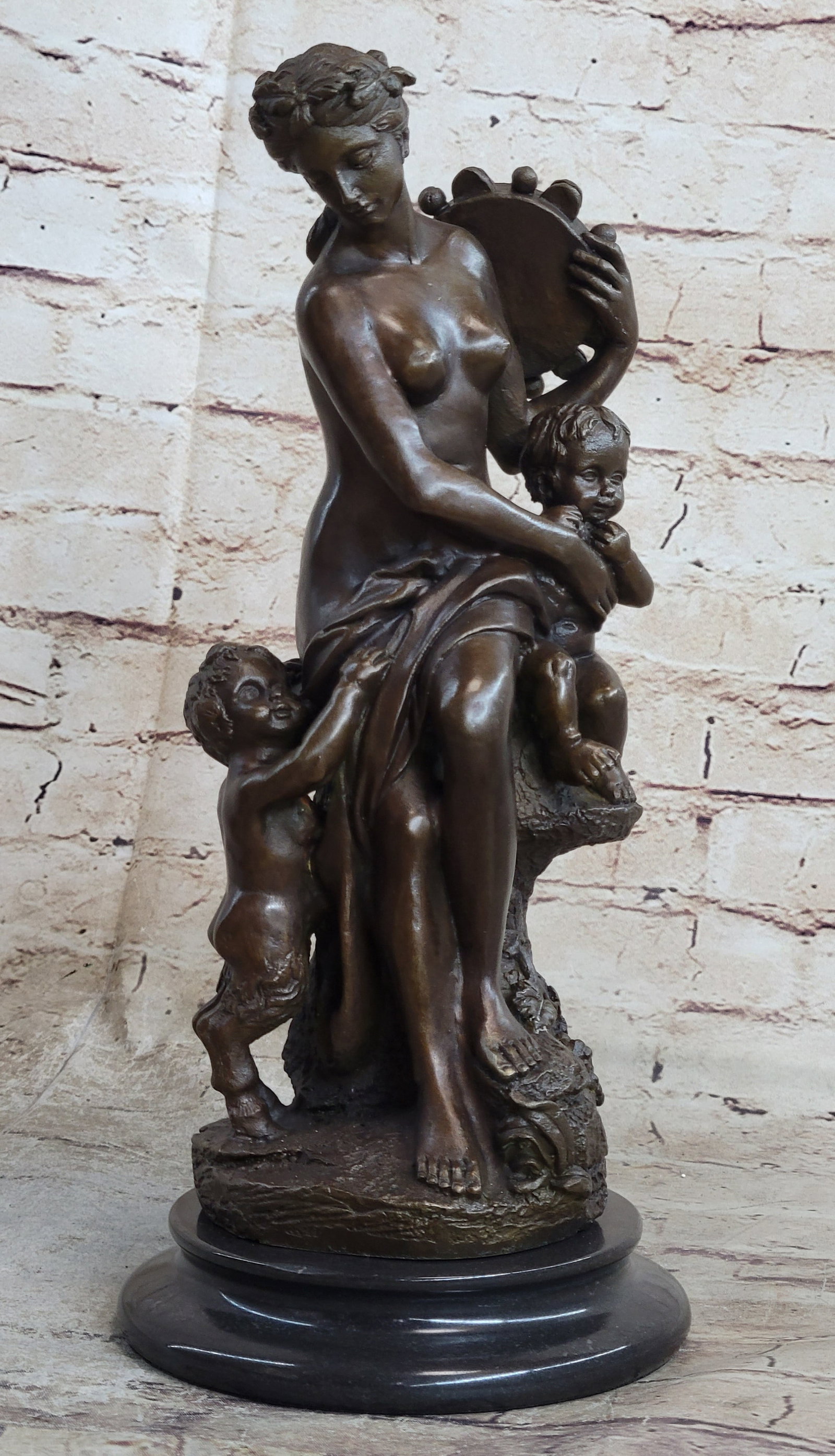 Woman with Cherubs Statue: The bronze sculpture, rich in Greek mythological lore, shows a beautiful nude maiden and two child figures. She sits upon a stone with a fabric robe twisting around her lower torso. Her hair is entwin