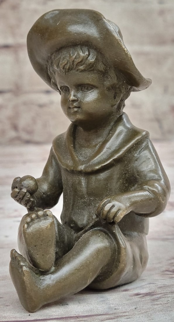 Boy Bronze Sculpture: Dressed in a sun hat revealing his neatly coiffed wavy hair, this young boy sits down with his legs folded. He holds a ball in his hands, as he is most likely taking a break from his play. He stares a