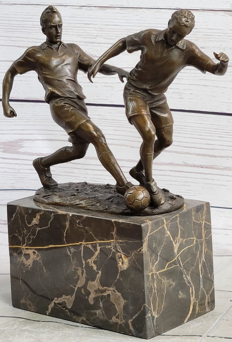 Two Soccer Player Bronze Sculpture: They are legends on the field. Two soccer players tumble down the grassy field, kicking, and dodging along the way. They are swift on their feet. One nears the goalpost and lunges one leg forward, pro