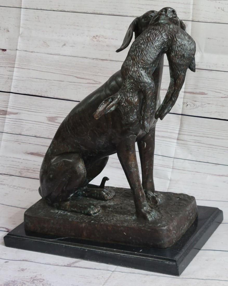 Hound Dog Hunter Bronze Sculpture: This sculpture depicts a dog, possibly a hound, with a rabbit gripped in his mouth. The hound is slim and his ribs are pronounced. His tail wraps around him as he sits. His eyes gaze up to the right, 