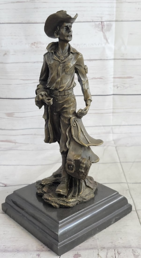 Old Cowboy Holding his Saddle Bronze Sculpture: This is a sculpture of a classic cowboy. He wears his cowboy hat and wearing a necklace around his neck. His jacket has hanging tassels and holding his saddle in his left hand. On his feet, she wears