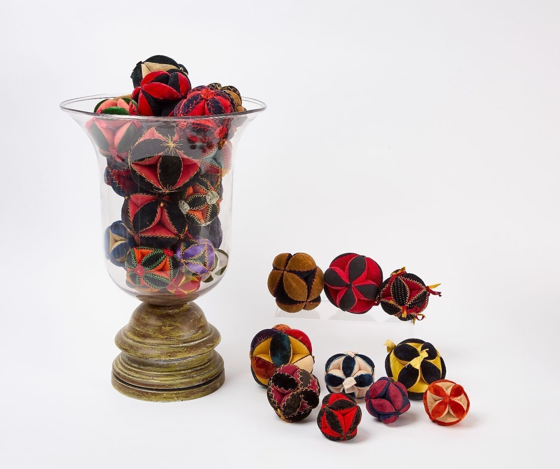 Large Group of Folk Puzzle Ball Pincushions in Glass Vase: Group of folk puzzle ball pincushions in various velvet, silk, and grosgrain fabrics, several with embroidered stitching, ribbon ties, or metal bead trim, arranged in a large glass hurricane/urn vase