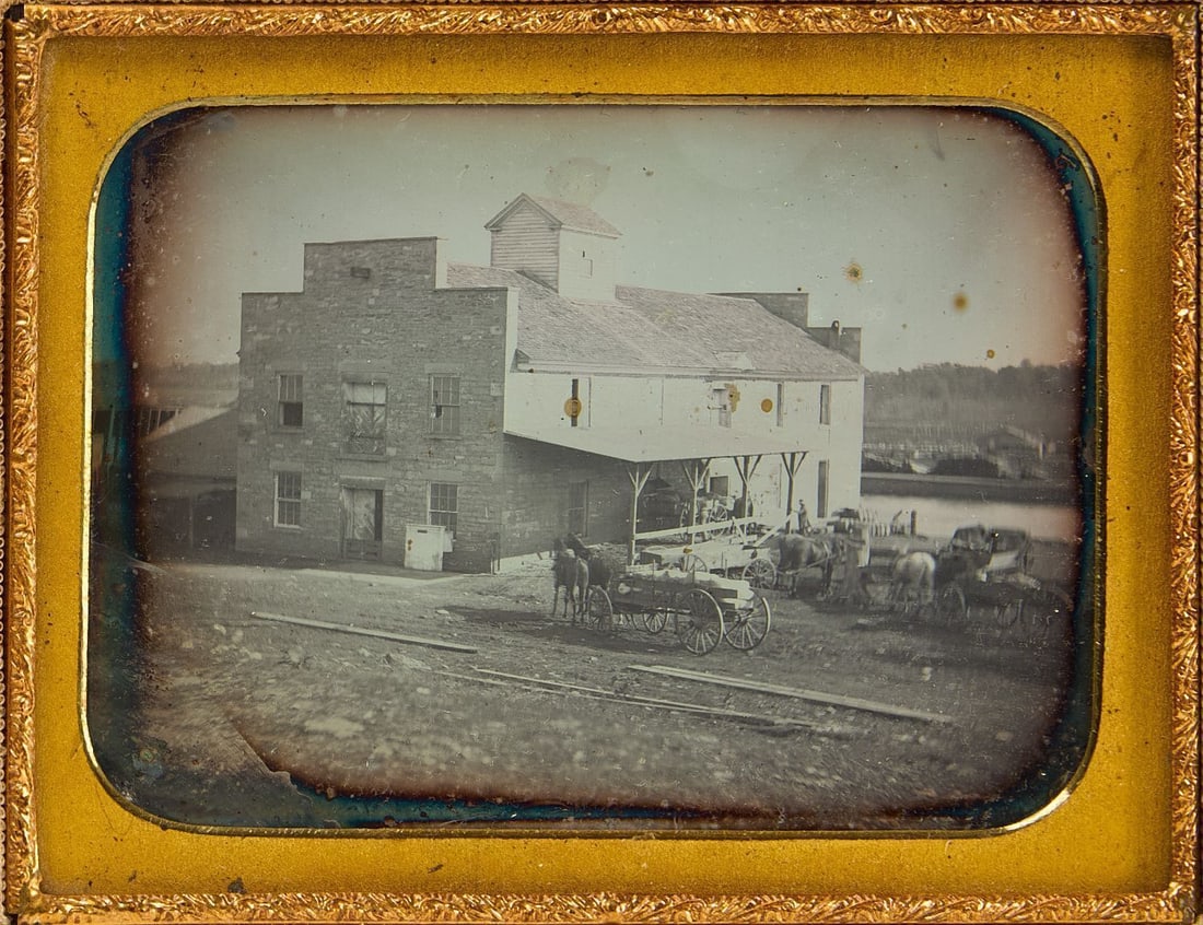 Daguerreotype Exterior View of a Mill: Quarter plate daguerreotype showing an exterior view of a building (likely a mill or factory), with horse-drawn wagons to the side. In the background appears to be a canal or river. Housed in a full