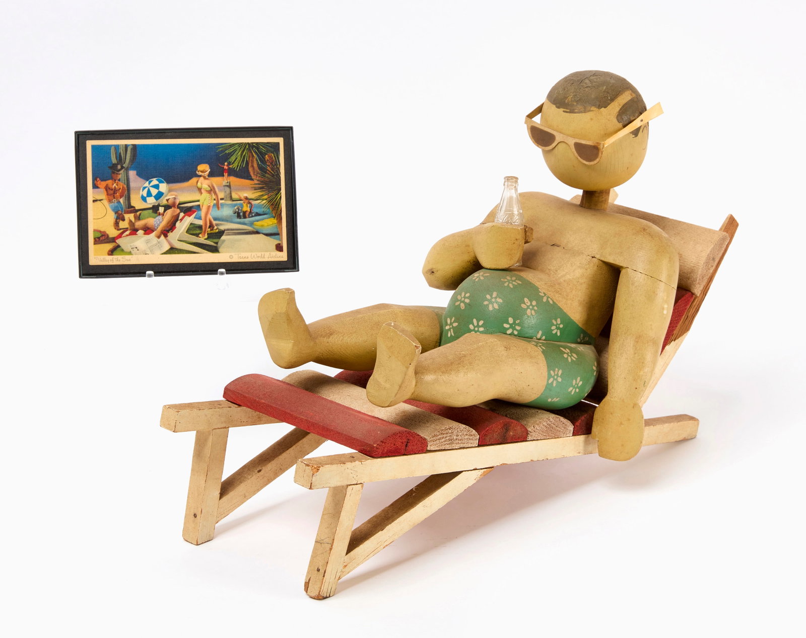 Trans World Airlines Carved Figure Display: Trans World Airlines original advertising display. Carved figure of a man in a swimsuit in a reclining chair holding a Coca Cola bottle. Carved and paint wood. Together with a framed postcard in