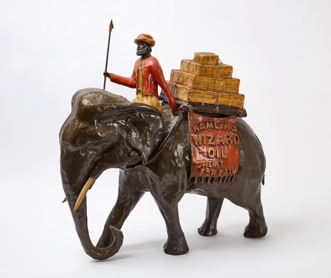 Hamlins Wizard Oil Elephant Countertop Display: Hamlins Wizard Oil countertop display in the figure of a merchant atop an elephant with packages on the elephant's back. Paper mache composition. American, circa 1920. H 28-3/4 in. W 26 in. D