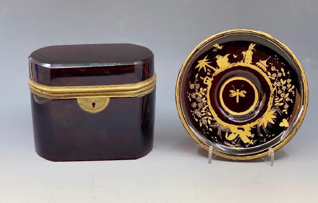 Ruby Flashed Glass Box and Ruby Chinoiserie Saucer: Squared oval box in ruby flashed glass, together with a saucer decorated with gilded chinoiseries. 4.5 in. w. x 5.5 in. h. Condition: Box with small chip to front lower corner, norma