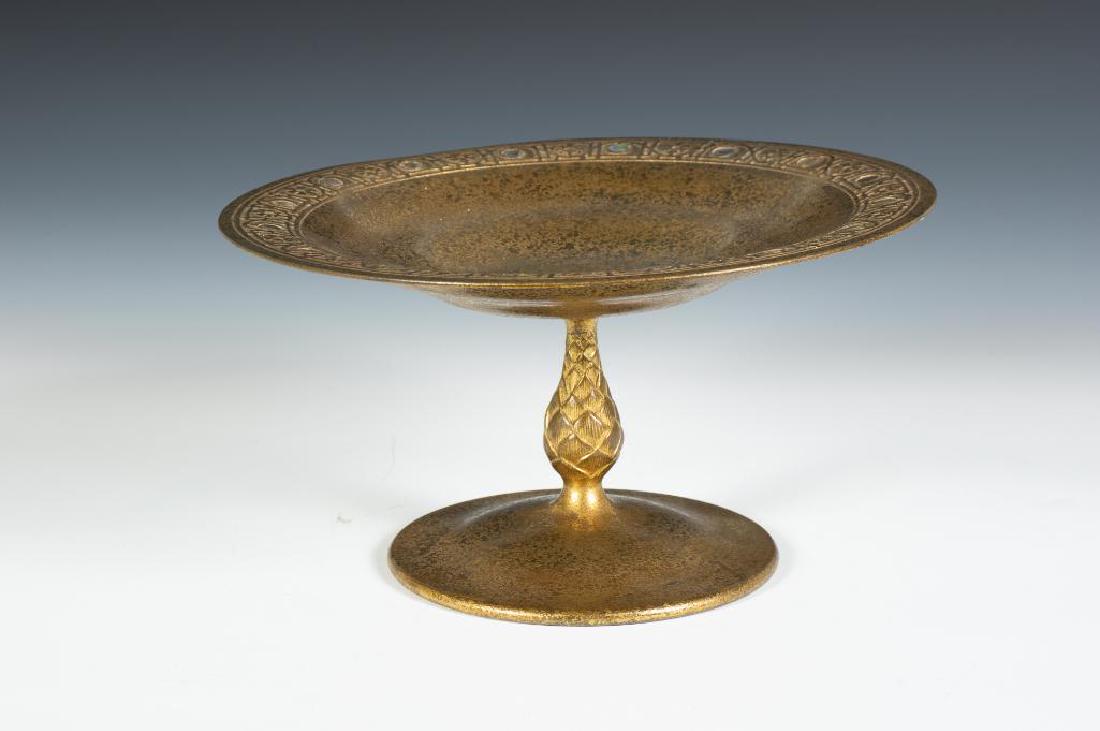 A Tiffany Bronze and Abalone Compote: The circular dish with a stylized floral border, set with abalone disks, raised on a seed pod standard and circular foot, stamped Tiffany Studios New York, 1664. 9.75 in. dia. Condition: Slightly bent