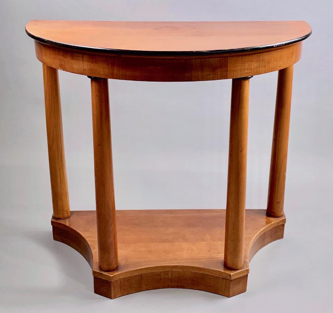 Beidermeier Demilune Console Table, c.1815: In pearwood (?) the demilune top with ebonized edge, raised on tapering columnar legs with ebonized capitals, all joined by a shaped stretcher base. 30 x 33.5 x 16 in. Condition: Minor vene