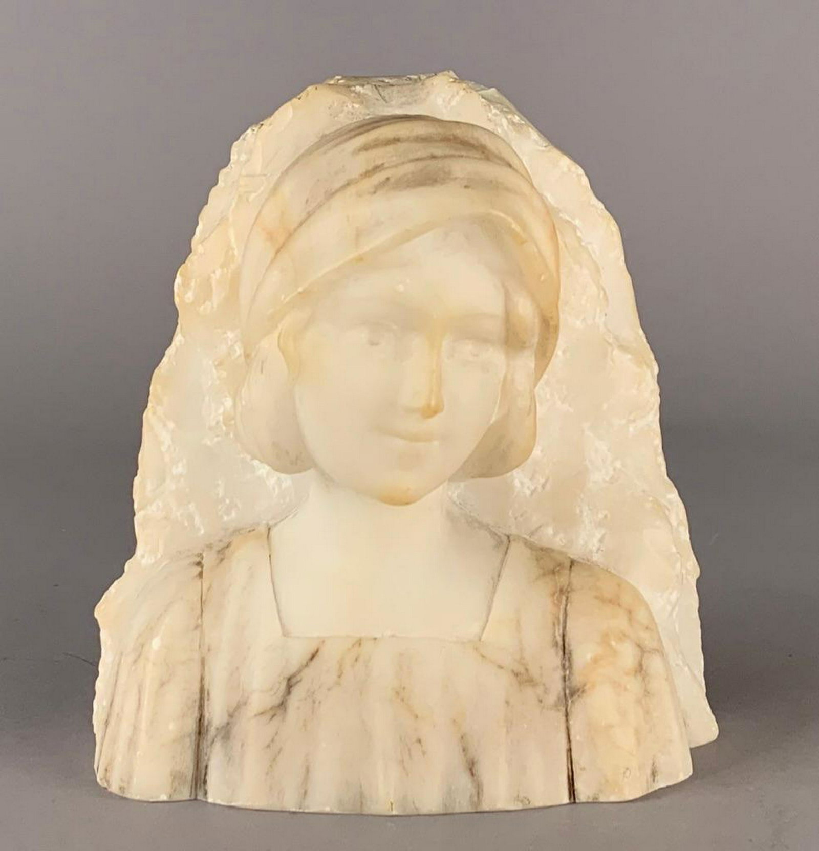 Alabaster Bust of a Young Girl: Early 20th C., bust length depiction of a young girl wearing bonnet in white and veined alabaster. 6.5 x 5 in. Condition: Wear and surface grime with some small flakes.