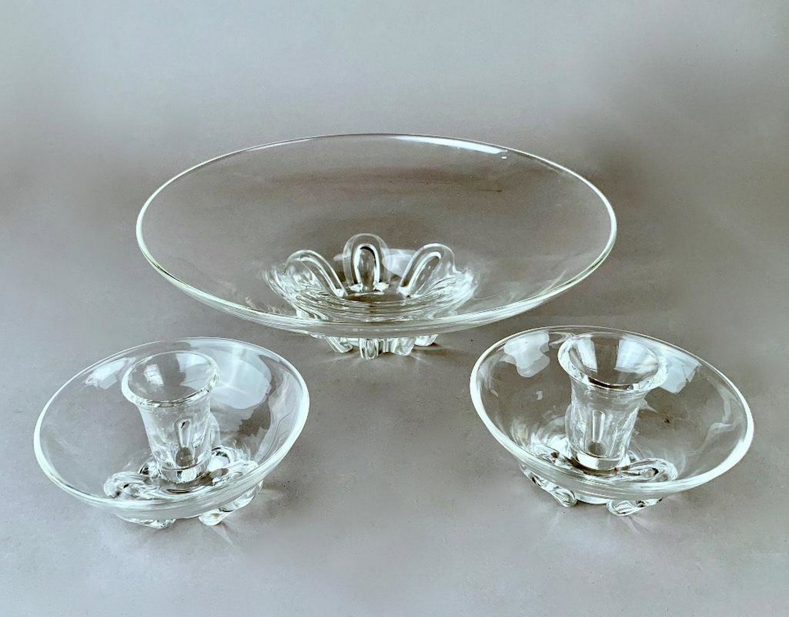 Steuben Three Piece Garniture: In colorless glass, comprising center bowl and two candlesticks, each signed. 12.75 in.dia. of bowl 5.5 in. dia. of candlesticks Condition: One candlestick bobeche ground. A few small chips to foot of