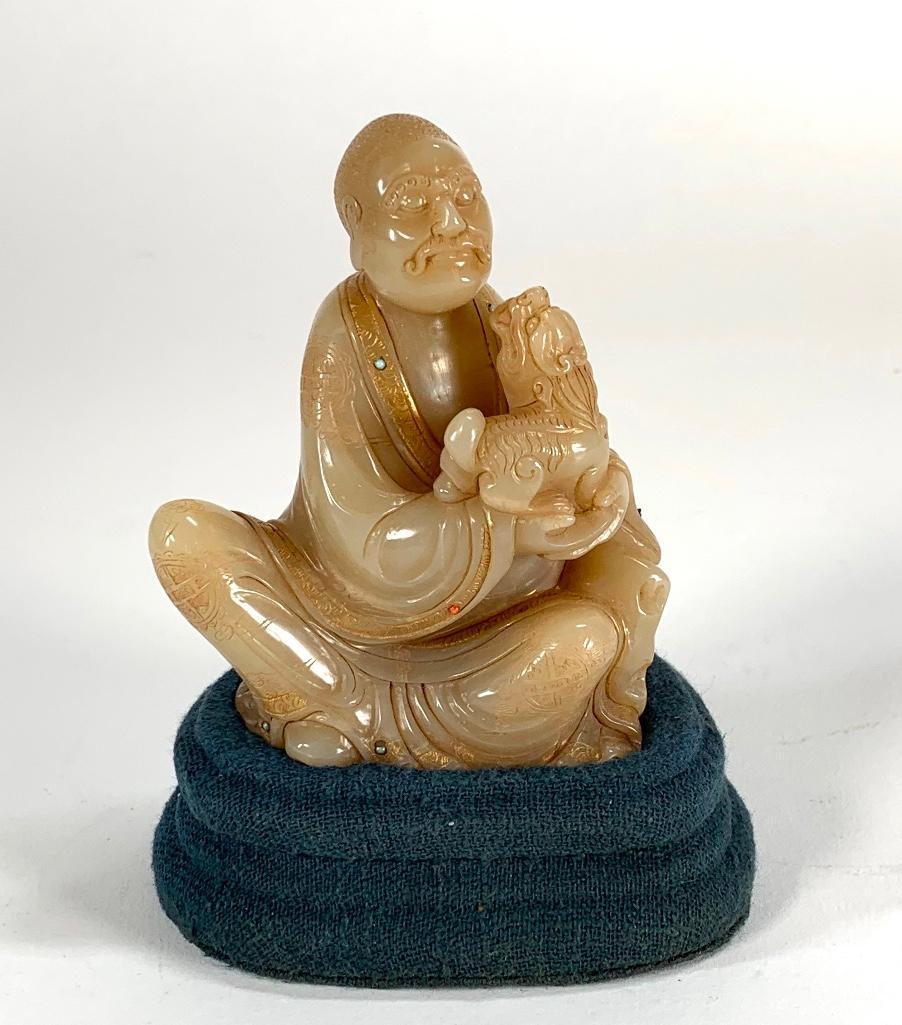Chinese Carved Soapstone Shoushan Lohan: Chinese Carved Soapstone Shoushan Lohan, nicely carved seated figure with dog, the Lohan's robes with gilded accents and set with small stones, in fitted case with stand. 4 x 3 in. figure Condition: S