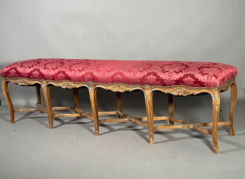Louis XV Style Beechwood Banquette: Louis XV Style Beechwood Banquette, shaped long form on ten carved cabriole legs joined by stretchers, upholstered in red damask Dimensions: 23 x 81 x 18 in.d. Condition: Very good.