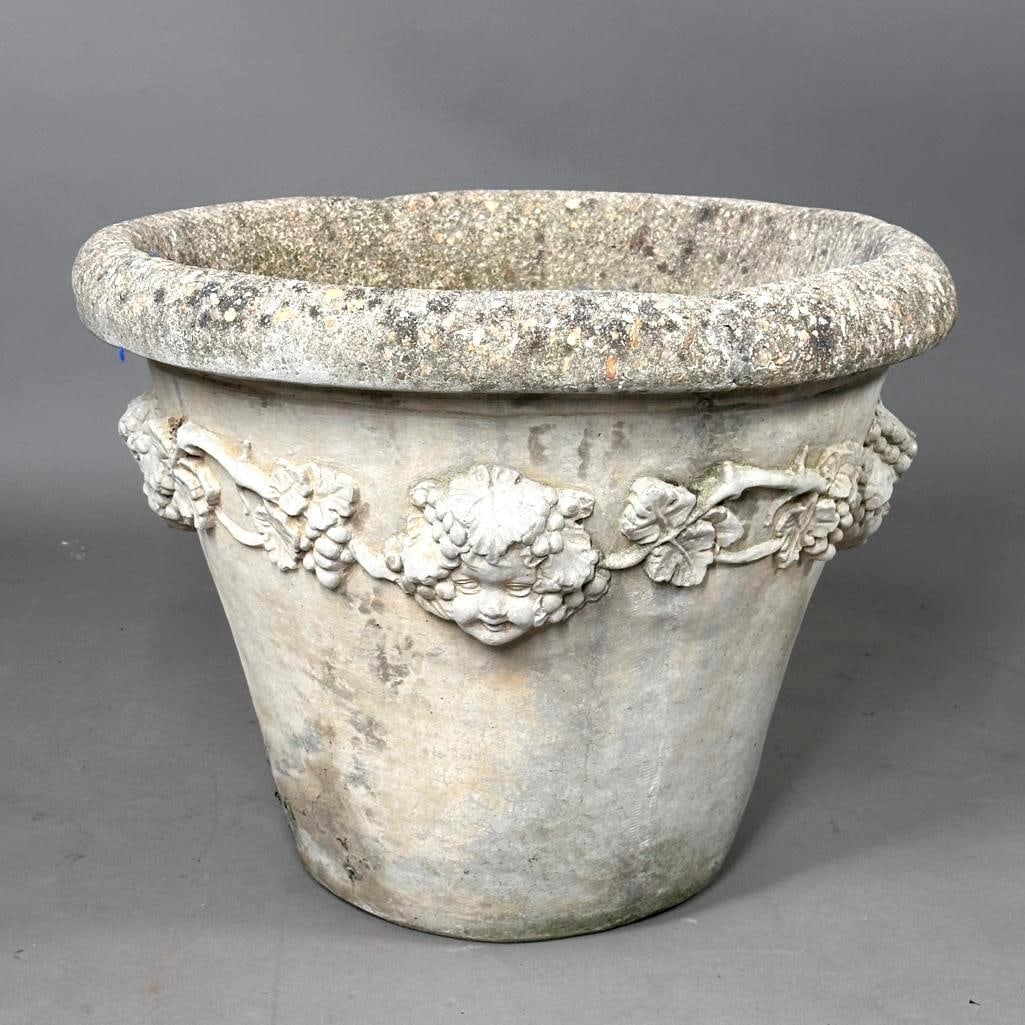 A Large Concrete Garden Planter Cast with Cheubs: A large cast concrete garden planter, modern, tapered conical form with foldover rim, the body cast around the form with cherub faces and fruiting grapevines. With three pot feet. Dimensions: 18.5