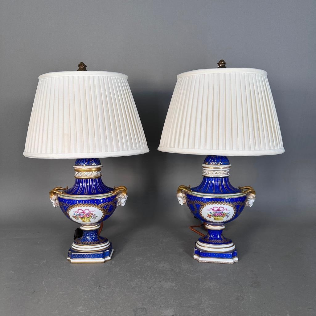 A Pair of Sevres Style Porcelain Boudoir Lamps: A Pair of Sevres Style Porcelain Boudoir Lamps, Modern. Covered vase form with ram's head handles, decorated with a floral basket reserved on a cobalt blue and gilded ground, on a stepped base.