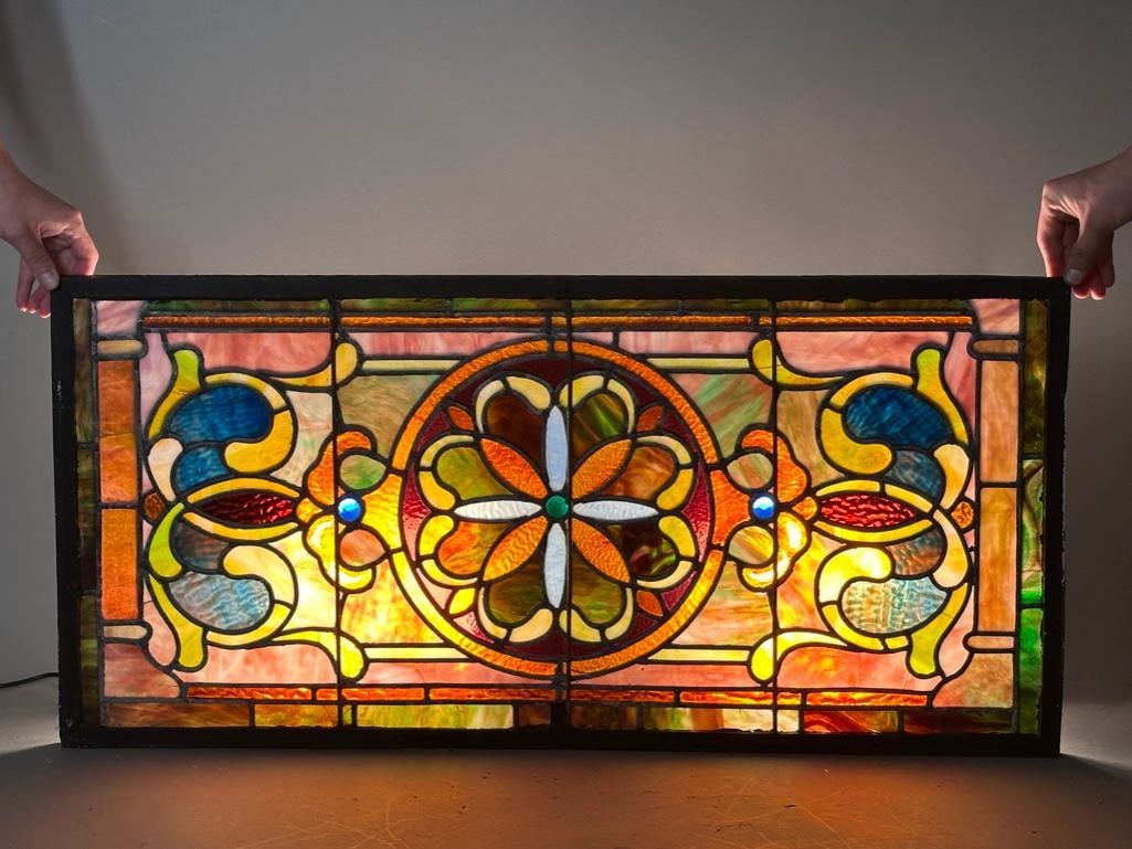 Leaded Glass Window: Leaded Glass Window, the rectangular form centered by a stylized flower form flanked by scrolls, in colorful striated and textured glass, in wrought iron frame. Dimensions: 18.5 x 38.5 in. Overall.