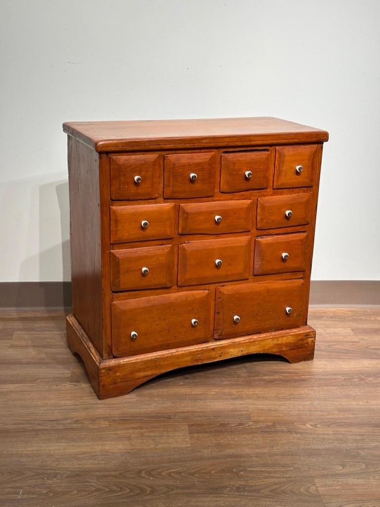 American Walnut Apothecary Chest: Pine cabinet with twelve drawers, white porcelain knobs, on open-carved base, 30 x 28.5 x 14.5 in. Condition: very good, stains insides drawers.