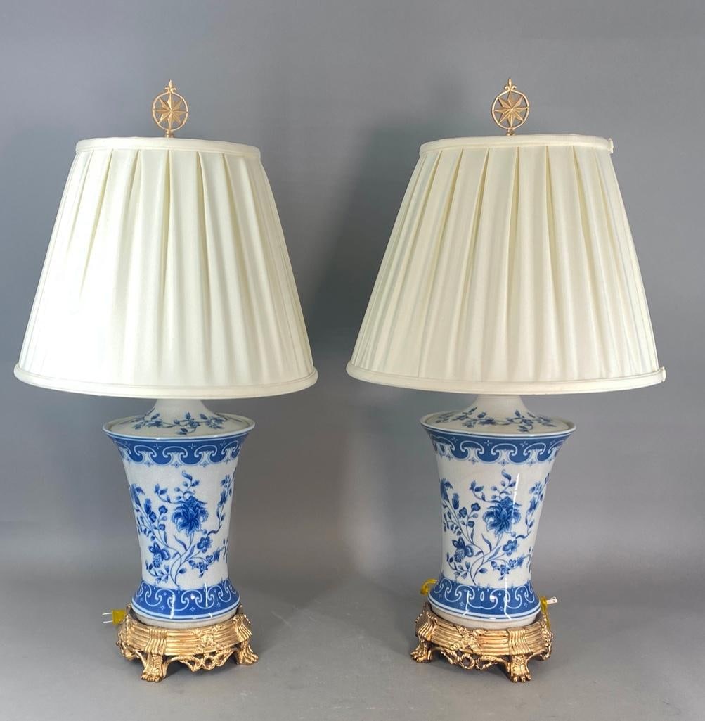 Pair of Chinoiserie Style Porcelain Lamps: White porcelain with a crackle-glaze, printed blue decoration with Iris flowers within a blue border of lappets. Mounted in a gilt metal base, electrified, one light each, with matching finials and sh