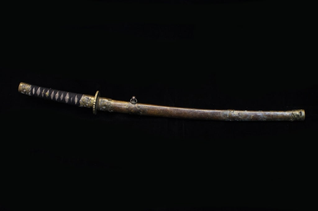 Japanese wakizashi Katana Sword: Scabbard (saya): The saya is made of metal, decorated with embossed motifs, including a detailed lion-dog (shishi) figure and other floral or symbolic designs. The patina suggests significant age and 