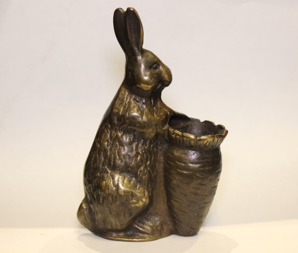 bronze sculpture of a rabbit holding a basket: Bronze sculpture of a rabbit holding a basket. The rabbit depicts a seated rabbit upright on its haunches, with both front paws resting on the rim of a woven-style basket. The basket is integrated int