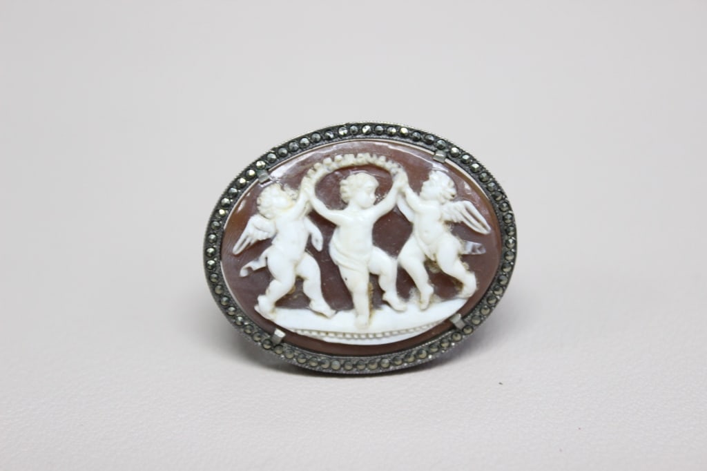 Vintage Cameo Brooch Depicting Three Figures: vintage cameo brooch depicting three figures, commonly identified as the Three Graces or putti (cherubs), set in a silver frame adorned with marcasite stones.Dimension: 3cm x 3.5cm