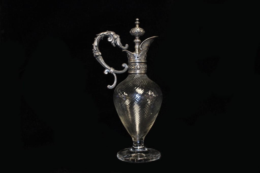 Antique Victorian Claret Jug: Dimension: 8"H. The clear glass body features a distinctive spiral-moulded or "thread glass" pattern that tapers into a pedestal foot. The neck, spout, and ornate handle are made of