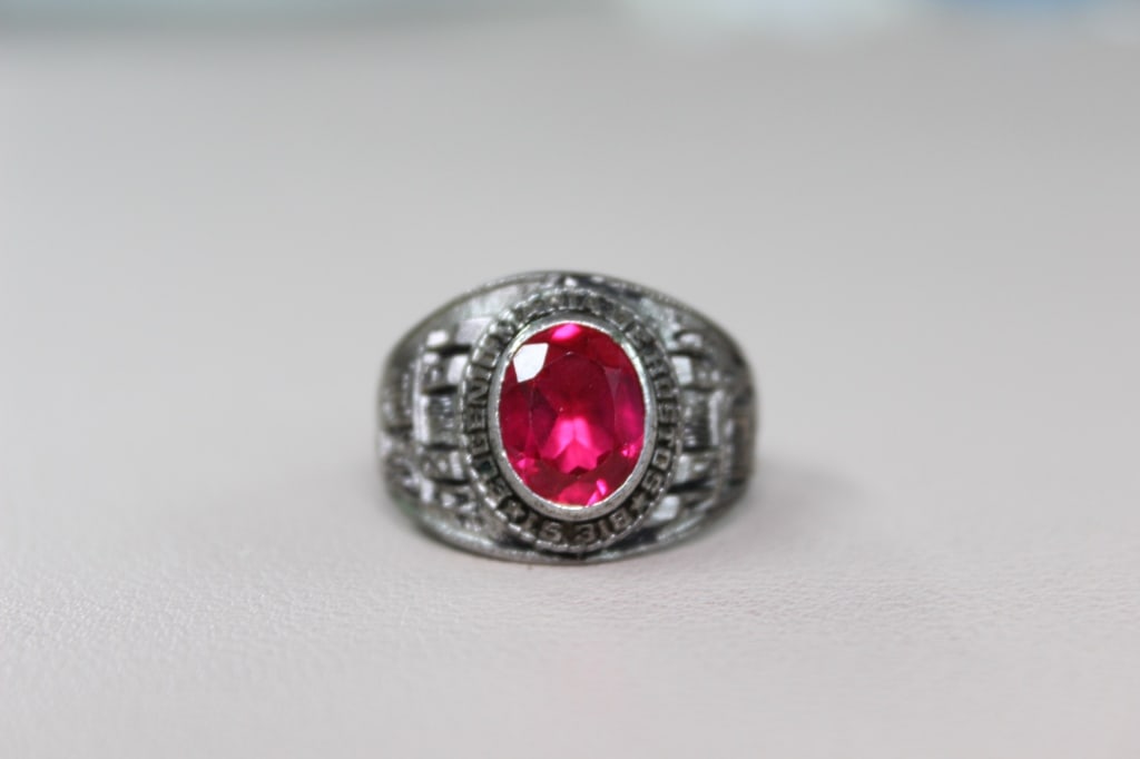 Faceted Synthetic Garnet Ring: The ring is designed in a traditional signet style, a common feature of high school and college class rings. The central red stone is set within a metal bezel that reads "CIKELIGEN" around its 