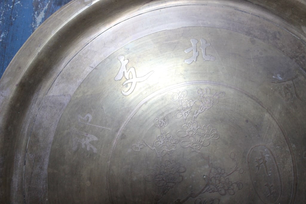 Antique Asian Circular Brass Platter: The center of the dish features a hand-etched, detailed illustration of blossoming floral branches, appearing to be plum or cherry blossoms. Flanking the central circle are larger, fluidly engraved ch