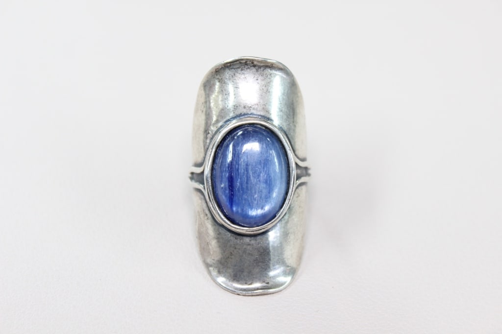 Bohemian-style Blue Kyanite Gemstone Ring: large, bohemian-style ring featuring a prominent, oval-shaped blue kyanite gemstone set in a simple bezel setting. The bold, chunky design creates a strong statement piece. The ring has an elongated,