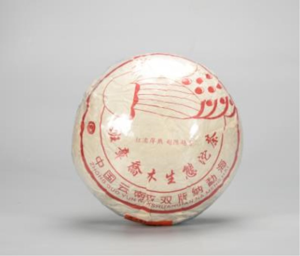 Chinese Banzhang Qiaomu Pu-erh tea cake: Dimension: 1800g The white paper wrapper is printed with distinctive red graphics across the upper half, featuring an abstract traditional lantern or basket motif flanked by stylized decorative dots.