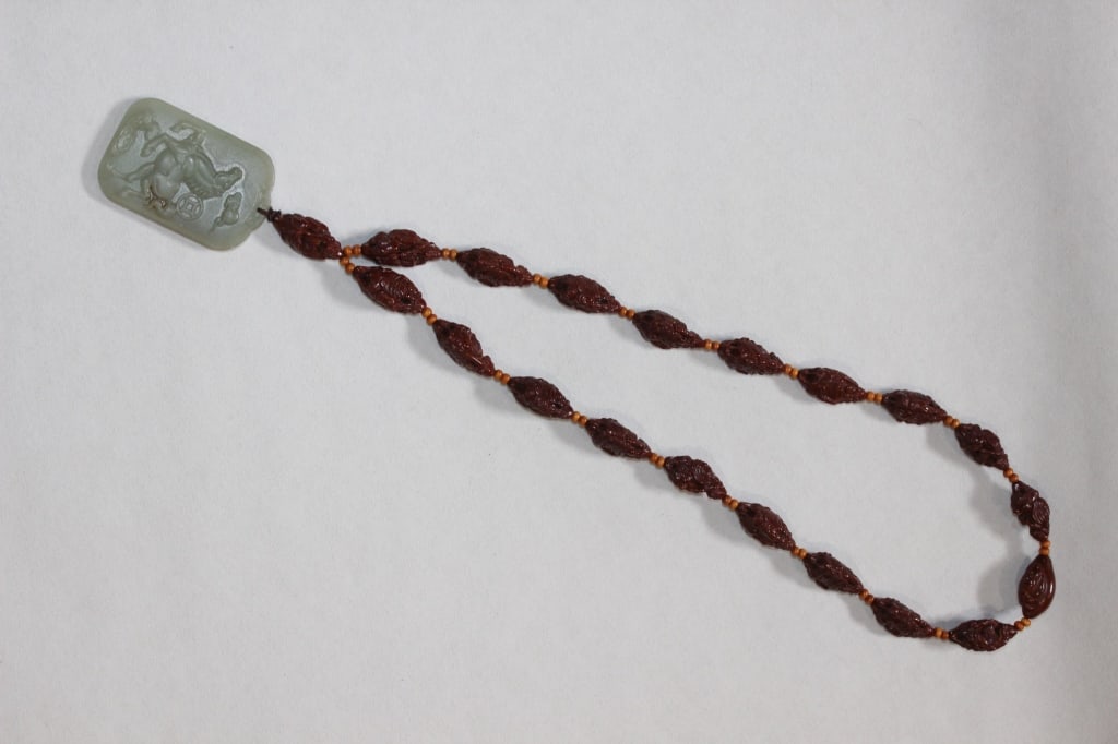 Chinese Carved Olive Pit Necklace w Jade Plaque: The necklace is composed of olive pits that have been meticulously hand-carved into pointed, oblong beads.The face of the jade plaque features a low-relief engraving, which appears to depict a