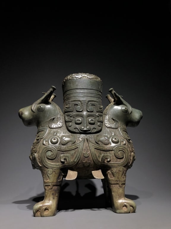 Chinese archaic Bronze ritual wine vessel: Dimension: 35cm w x 12.5x 37cm H. The vessel features two stylized rams standing back-to-back, sharing a conjoined four-legged torso. Their necks and sculpted heads protrude outward dynamically on opp