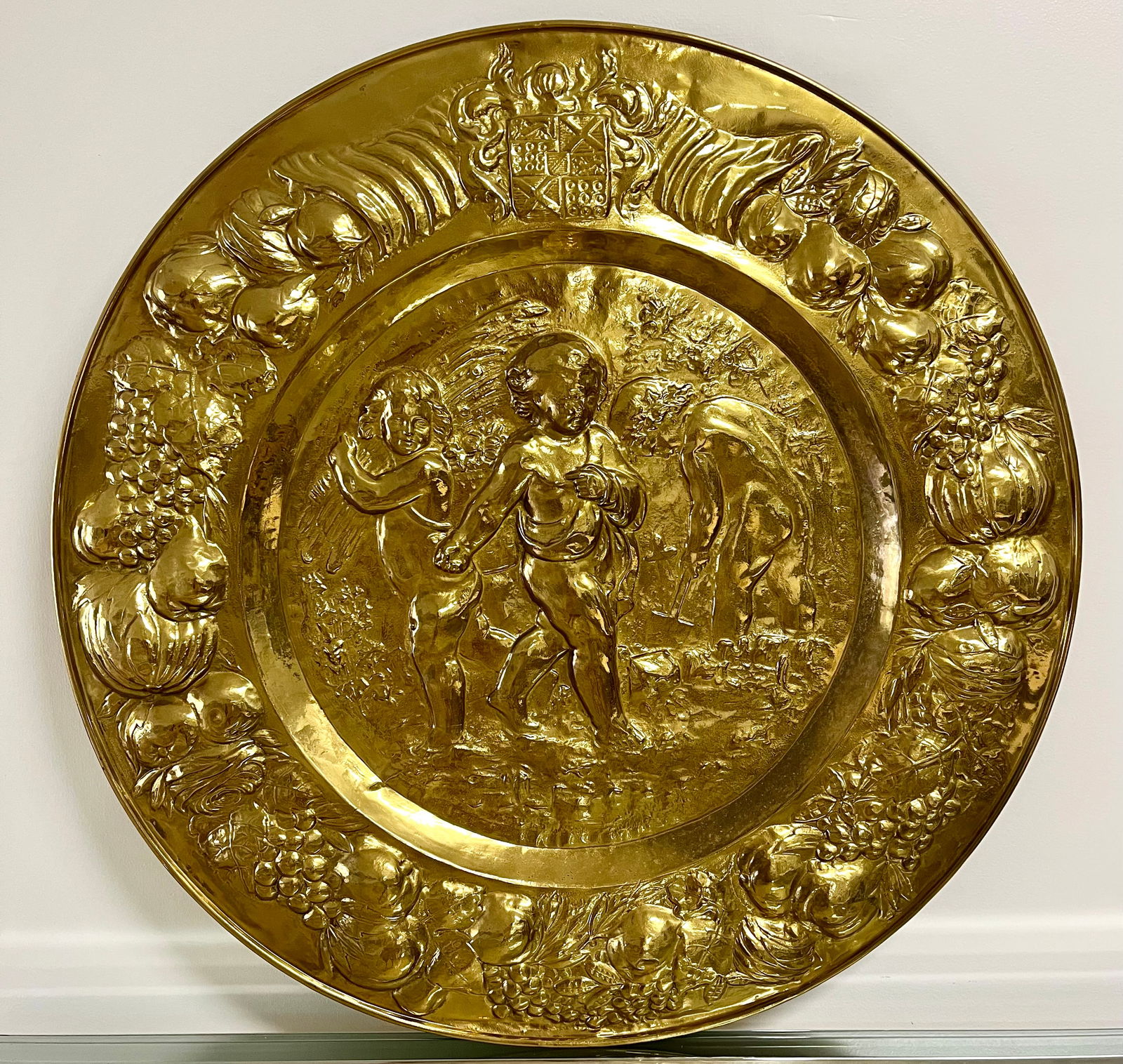 Large 29?? Ornate Brass Wall Charger | Repousee Cherubs & Fruit Rococo Style: Striking 29” repoussé brass wall charger featuring cherubs, fruit garlands, and a heraldic crest in ornate Renaissance Revival style. This grand decorative piece is richly embossed and