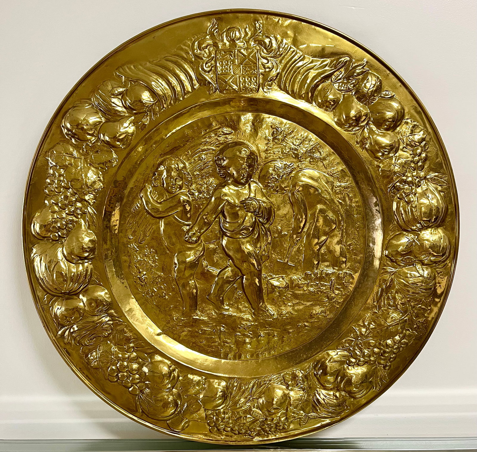 Large 29?? Ornate Brass Wall Charger | Repousee Cherubs & Fruit Rococo Style: Striking 29” repoussé brass wall charger featuring cherubs, fruit garlands, and a heraldic crest in ornate Renaissance Revival style. This grand decorative piece is richly embossed and finished