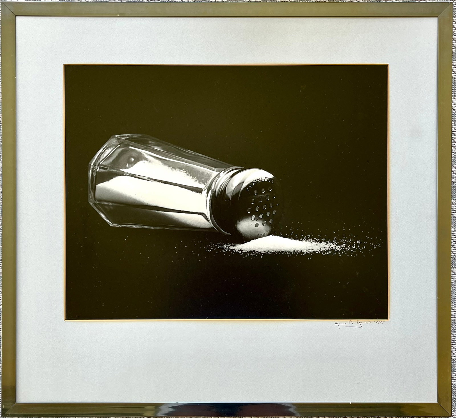 1979 Black & White Salt Shaker Photograph Signed: Original black and white photograph dated 1979, featuring a stainless steel salt shaker tipped on its side, spilling salt across a dark background in striking contrast. Professionally custom framed an