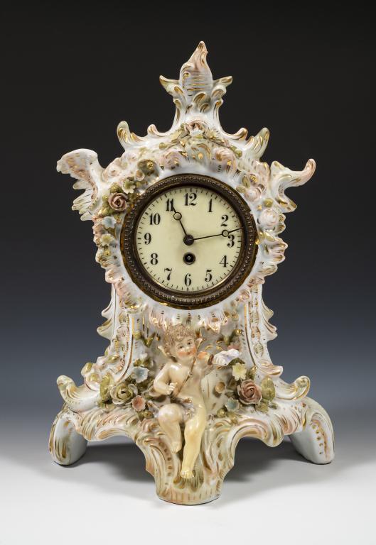 Hamburg American Clock Co. Figural Porcelain Clock: Item/Description: Porcelain clock case embellished with applied hand-painted flowers and garnished with gilt and scrolled accents. Porcelain cherub figurine sits center below porcelain clock face with
