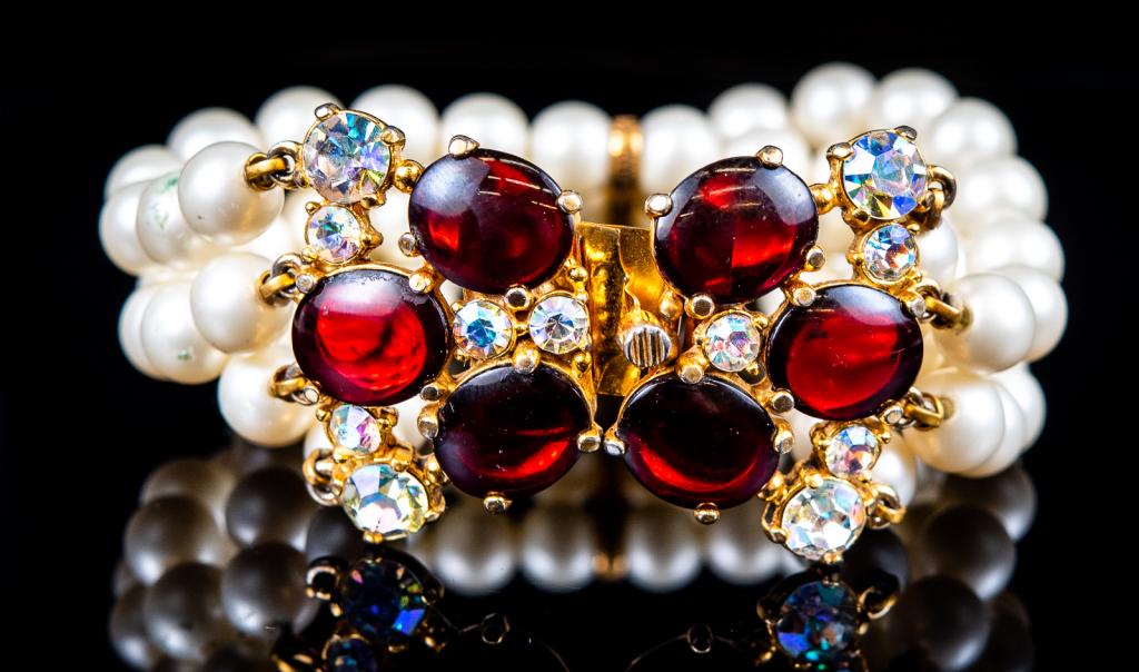 Elsa Schiaparelli Triple Strand Pearl Bracelet: Item/Description: Rare bracelet by the iconic Italian fashion designer. Three strands of lustrous white faux pearls threaded through antique goldtone bar; jeweled floral push-in clasp of cabochon glas