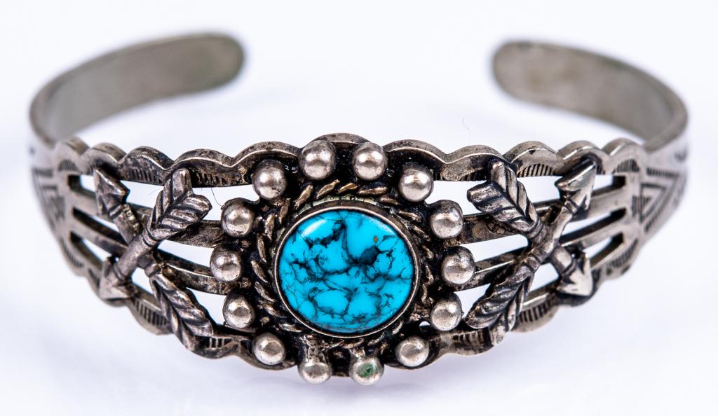 Native American Silver Tone Turquoise Cuff: Item/Description: Ornate silver tone cuff bracelet with deep blue turquoise florette medallion, striated open band with crossed arrows, engraved Native American symbols. No visible hallmarks. Material