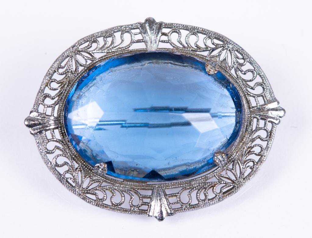 Art Deco Rhodium Plated Blue Glass Brooch: Item/Description: Exquisite Deco era antique brooch. Fashioned from rhodium plated metal and set with a cornflower blue glass faceted gemstone framed in a pierce-work filigree, open-backed setting. Ro